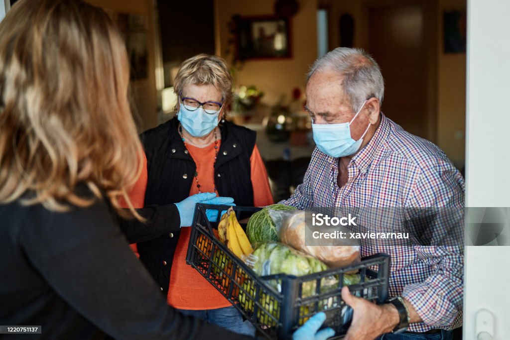 Grandchild delivers groceries to grandparents during pandemic at their home Senior couple on their 70s wearing a protective face mask picking up the groceries box that granddaughter is delivering in times of COVID-19, she is wearing mask and gloves. Senior Adult Stock Photo