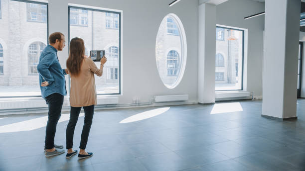 istock Young Hipster Man and Female Stand in an Empty White Office and Map it with an Augmented Reality Software on a Tablet. Sunlight Shines Through Big Windows. 1220765618