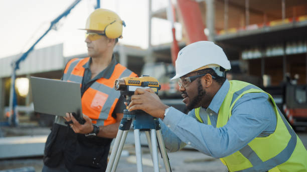 istock On the Commercial / Industrial Building Construction Site: Professional Engineer Surveyor Takes Measures with Theodolite, Worker Uses Laptop. In the Background Skyscraper Formwork Frames and Crane 1220765723
