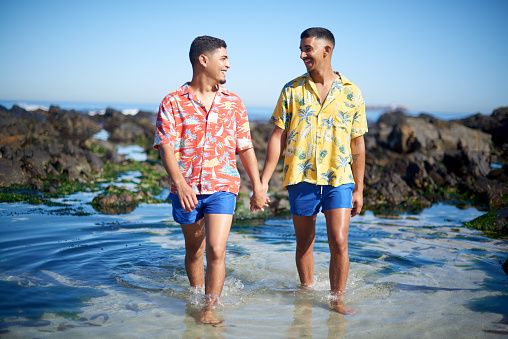 Full length shot of an affectionate gay couple holding hands while walking through a tidal pool at the beach