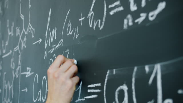 Close-up of male hand writing mathematics equations on chalkboard in class
