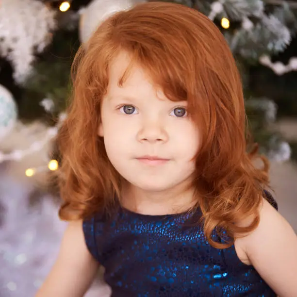 Christmas happy girl. Luxury attractive red hair child. Xmas miracle. Green tree, lights, bokeh.