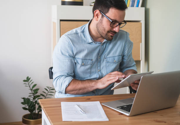 istock Handsome young man using digital tablet, sitting at a table with laptop and business papers stock photo 1220770339