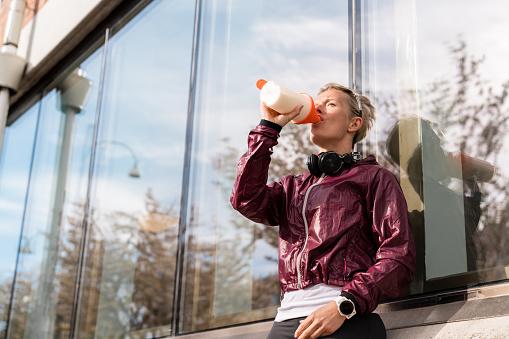 Woman exercising in the city. She is taking a break to drink a protein shake from a plastic bottle.