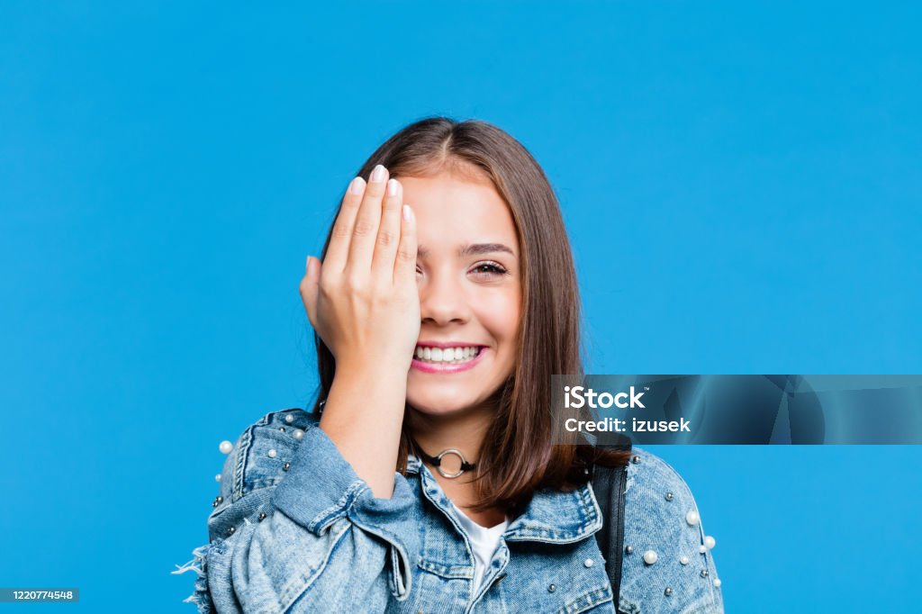 Cheerful pretty teenege girl on blue background Cute female high school student wearing oversized denim jacket and white t-shirt standing against blue background. Portrait of smiling teenager covering her eye with hand. Teenage Girls Stock Photo