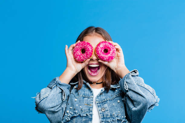 Excited teenage girl covering eyes with donuts Cute female high school student wearing oversized denim jacket and white t-shirt standing against blue background. Portrait of smiling teenager covering her eyes with donuts. double denim stock pictures, royalty-free photos & images