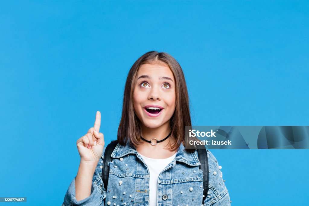 Happy teenge girl pointing with finger at copy space Cute female high school student wearing oversized denim jacket and white t-shirt standing against blue background. Portrait of surprised teenager looking up and pointing with index finger at copy space. Teenager Stock Photo