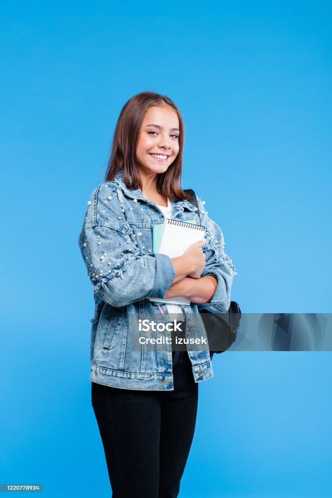 Portrait of female high school student holding notebooks Happy cute teenage girl wearing oversized denim jacket and black jeans holding notebooks. Smiling teenager standing against blue background. Girls Stock Photo