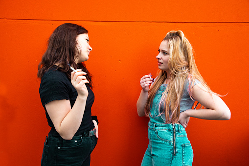Young woman, best friends, standing together in front of orange urban wall having a discussion about the problems they have right now with each other to find a solution for their conflict while smoking a cigarette together. Youth Culture Lifestyle Concept Shot.