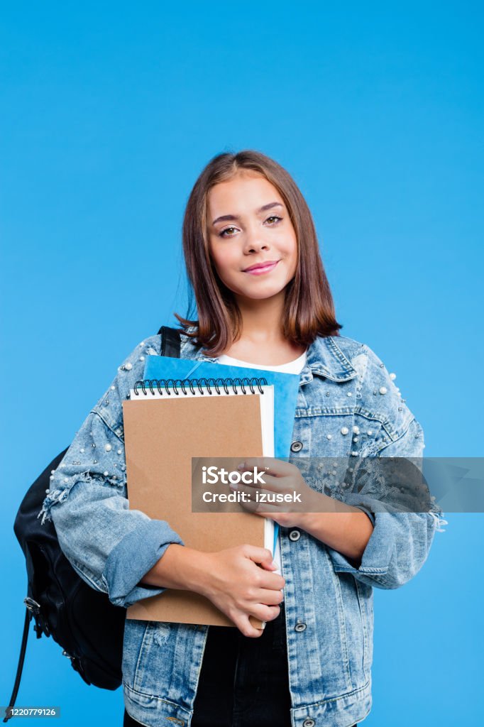 Portrait of female high school student holding notebooks Happy cute teenage girl wearing oversized denim jacket, black jeans and backpack holding notebooks. Smiling teenager standing against blue background. Student Stock Photo