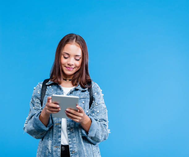 Portrait of female high school student using digital tablet Happy teenage girl wearing oversized denim jacket, white t-shirt and black jeans standing against blue background. Portrait of smiling teenager watching digital tablet. double denim stock pictures, royalty-free photos & images