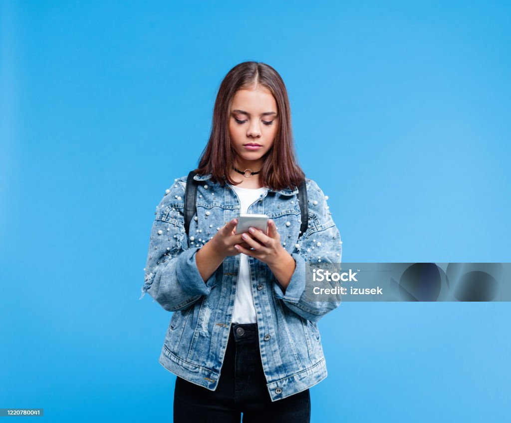 Portrait of female high school student using smart phone Teenage girl wearing oversized denim jacket, white t-shirt and black jeans standing against blue background. Portrait of teenager texting on mobile phone. Teenager Stock Photo