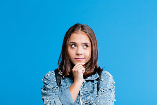 Cute female high school student wearing oversized denim jacket and white t-shirt standing against blue background. Portrait of pensive teenager with hand on chin.