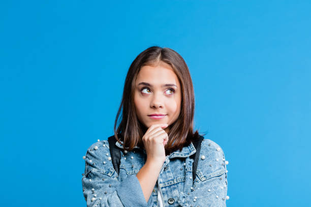 Portrait of pensive teenege girl Cute female high school student wearing oversized denim jacket and white t-shirt standing against blue background. Portrait of pensive teenager with hand on chin. rolling eyes stock pictures, royalty-free photos & images