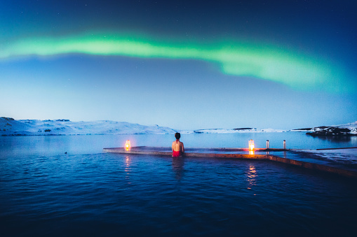 Woman in red swimsuit swimming in the hot thermal pool at the frozen lake looking at the dramatic bright Aurora dance and the snowy mountain peaks in East Iceland