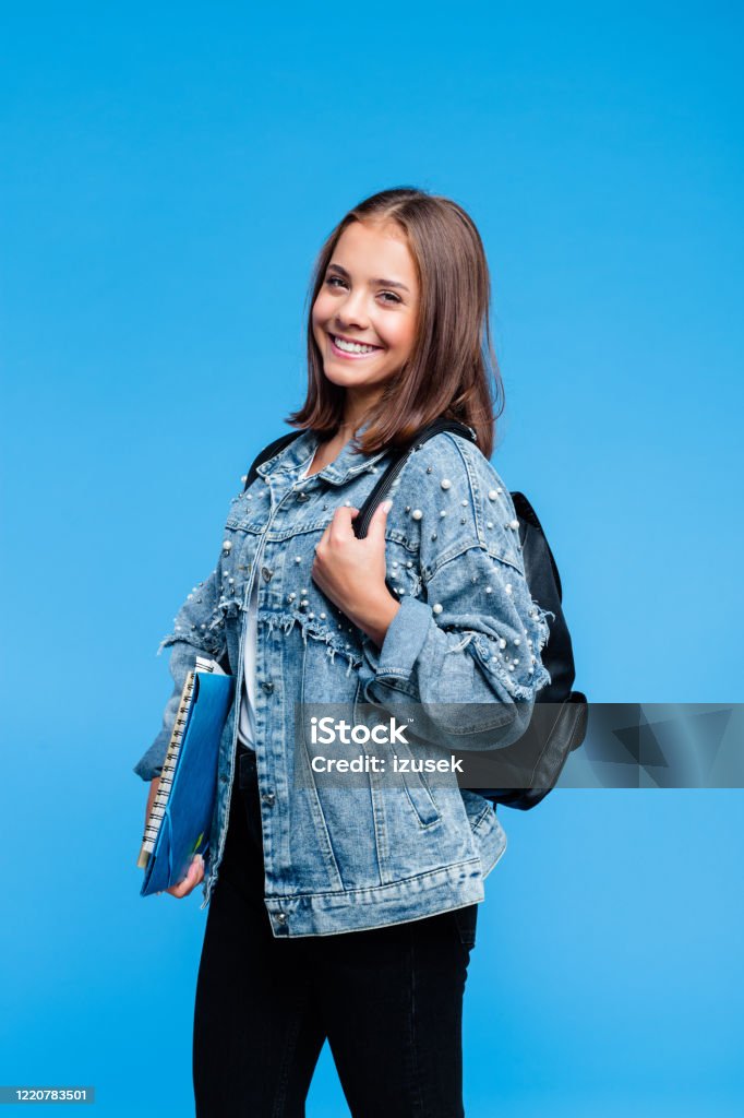 Portrait of pretty female high school student Happy teenage girl wearing oversized denim jacket, white t-shirt, black jeans and backpack standing against blue background. Pretty teenager holding books and notebooks. Student Stock Photo