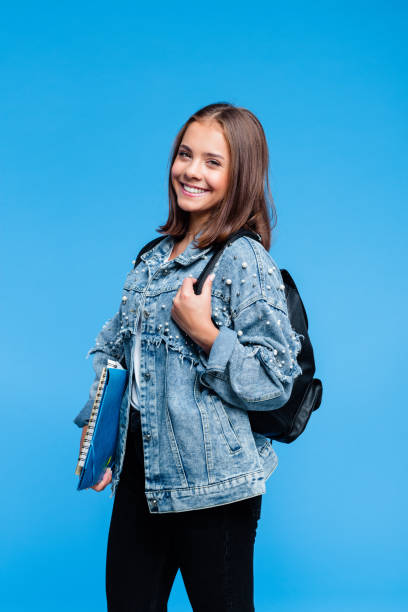 Portrait of pretty female high school student Happy teenage girl wearing oversized denim jacket, white t-shirt, black jeans and backpack standing against blue background. Pretty teenager holding books and notebooks. double denim stock pictures, royalty-free photos & images