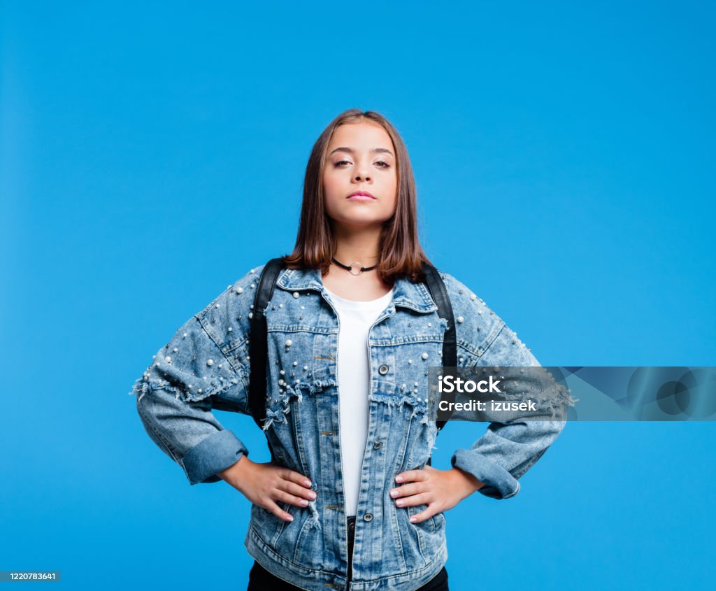 Portrait of super tennage girl Confident female high school student wearing oversized denim jacket and white t-shirt, standing against blue background. Portrait of power girl. Portrait Stock Photo