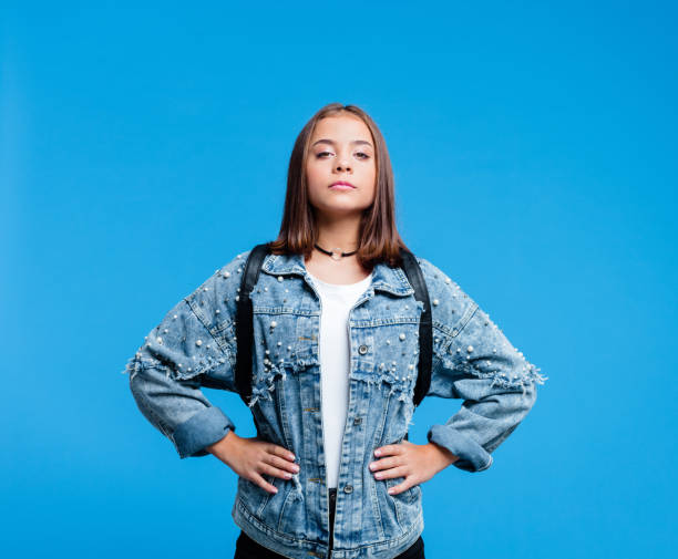 Portrait of super tennage girl Confident female high school student wearing oversized denim jacket and white t-shirt, standing against blue background. Portrait of power girl. physical position stock pictures, royalty-free photos & images