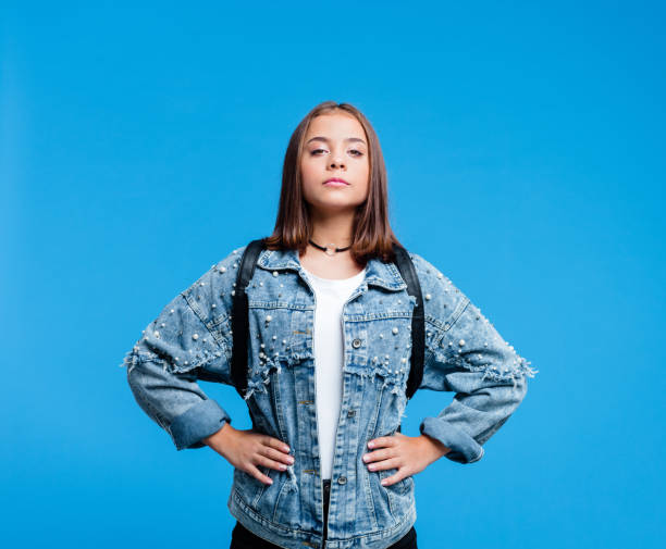 Portrait of super tennage girl Confident female high school student wearing oversized denim jacket and white t-shirt, standing against blue background. Portrait of power girl. child hero pose stock pictures, royalty-free photos & images