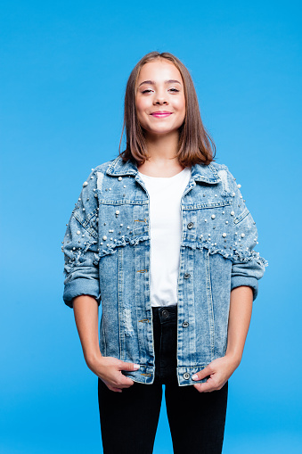 Portrait of teenage girl wearing oversized denim jacket, white t-shirt and black jeans standing against blue background. Portrait of smiling teenager.