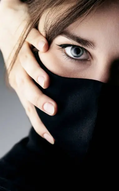 Photo of close up portrait of young beautiful elegant woman in black mask and hand clasped her face on studio background, computer hacker, spy girl, cyber security.