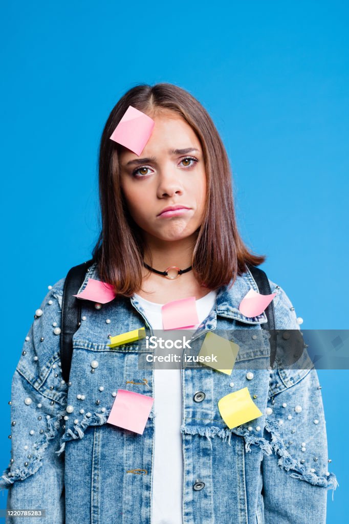 Worried teenager with adhesive note on forehead and jacket Displeased female high school student wearing oversized denim jacket and white t-shirt standing against blue background. Portrait of worried teenage girl with adhesive note on forehead and jacket. Reminder Stock Photo