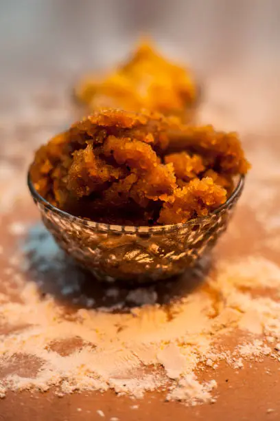 Close up of Indian Gujarati popular dish Atte ka sheera or Halwa-Karah parshad in a glass bowl on a brown surface with some spread wheat flour and some jaggery in a glass bowl. Vertical shot.