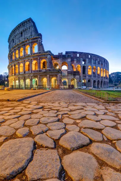 The illuminated Colosseum in Rome at dawn with big cobblestones in the foreground
