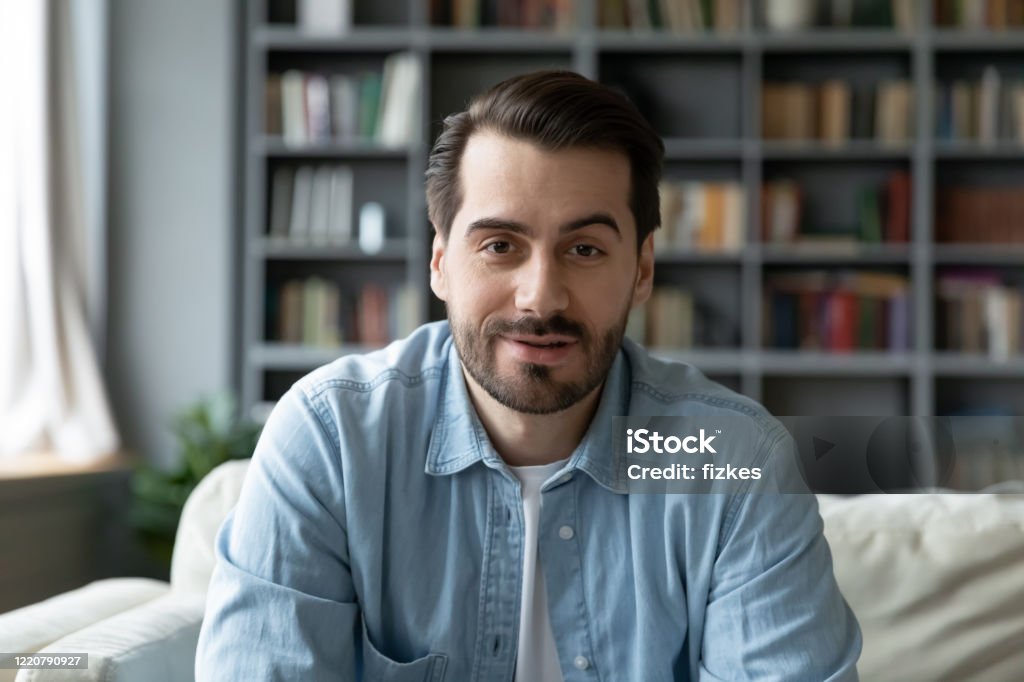 Headshot portrait millennial guy sit on sofa makes video call Head shot millennial guy sit on sofa in living room makes video call looks at camera, conversation by distant videocall, distance hiring job interview process, tutor and trainee study on-line concept Men Stock Photo