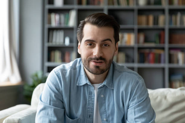 headshot portrait millennial guy sit on sofa makes video call - looking in camera imagens e fotografias de stock