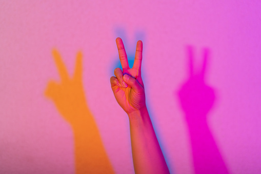 A close-up photo of a hand lit by neon lights doing peace sign.