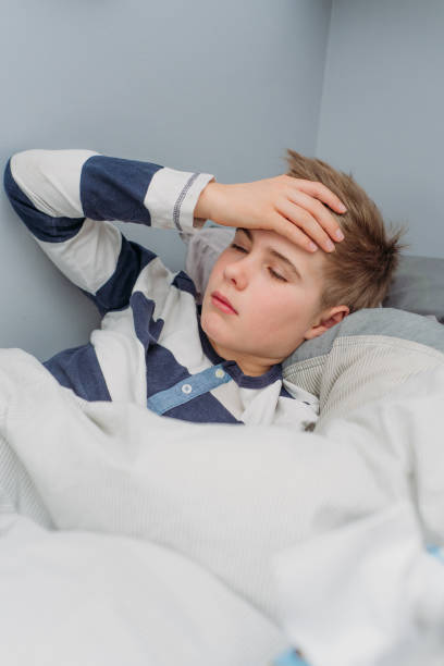 istock A 10 year old boy has a hand on his forehead and is lying ill in bed, which has white and grey cover. 1220798148
