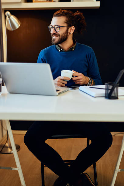 istock Young handsome bearded hipster sitting at his home office looking away and smiling. 1220806586