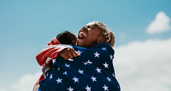 Excited american female athletes wrapped in national flag hugging each other after a winning the race. Team of USA female runner rejoicing a victory outdoors.