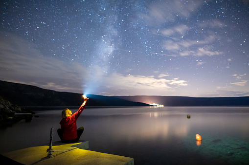Rear view of a blond woman shining a flashlight at the sky on seashore at night with the Milky Way visible above