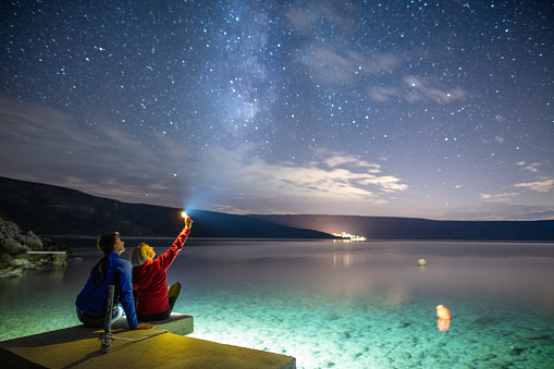 Rear view of a couple with a blond woman shining a flashlight at the sky on seashore at night with the Milky Way visible above