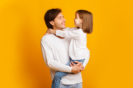 Cheerful father and little daughter posing together on yellow background, dad holding girl, looking at each other