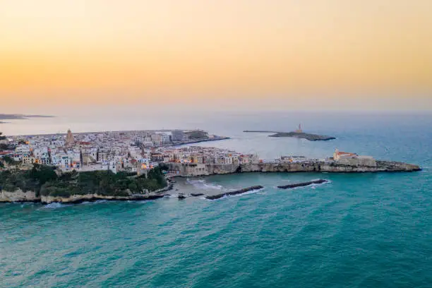 Aerial view of a coastal old town on a peninsula and cliffs along Adriatic Sea shore under a clear orange sky at sunset, Vieste, Puglia, Italy