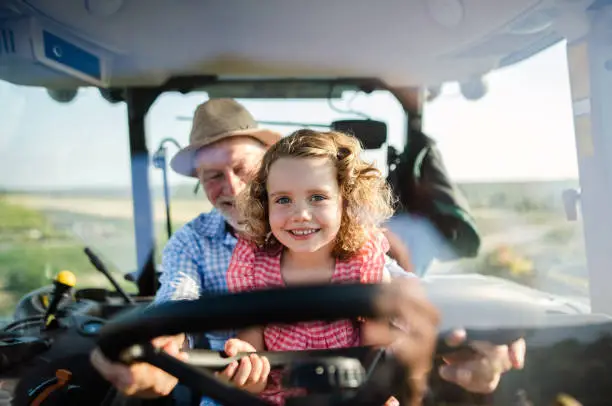 Photo of Senior farmer with small granddaughter sitting in tractor, driving.
