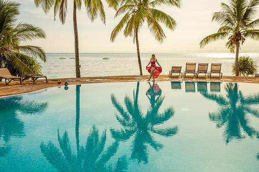 Female tourist in a red dress walking around a swimming pool surrounded by palm trees on a tropical vacation, Zanzibar, Tanzania