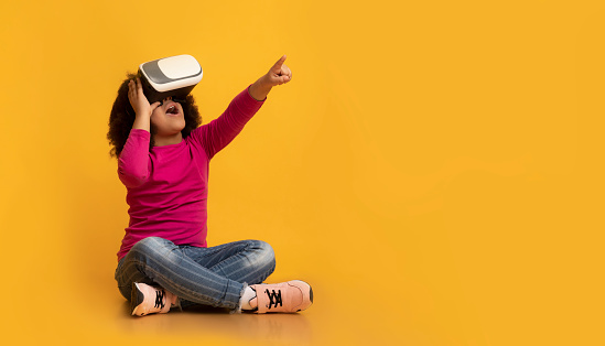 Little Afro Girl Wearing VR Headset, Experiencing Virtual Reality And Pointing Away While Sitting On Floor Over Yellow Background In Studio, Panorama