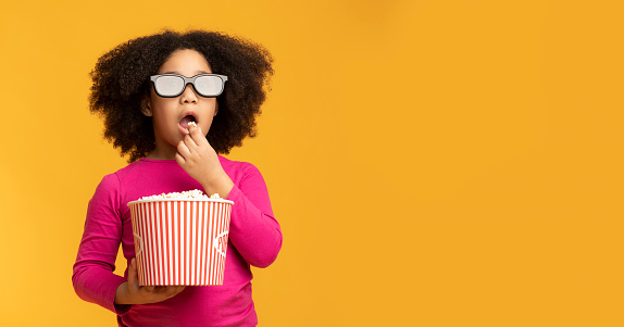 Kids cinema deals. Black little girl wearing 3D glasses and eating popcorn with dazed face expression, standing over yellow background, panorama