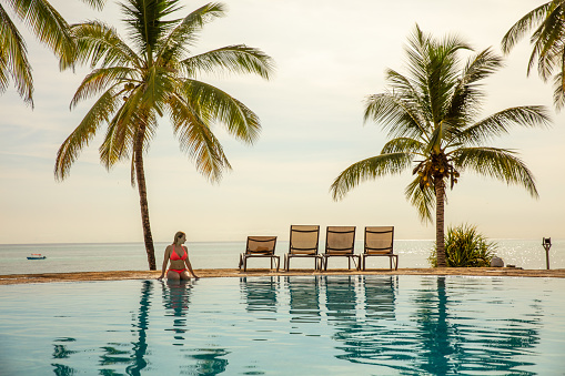 Young blonde woman in bikini sitting on pool edge at tropical sandy beach with palm trees at sunset, Zanzibar, Tanzania