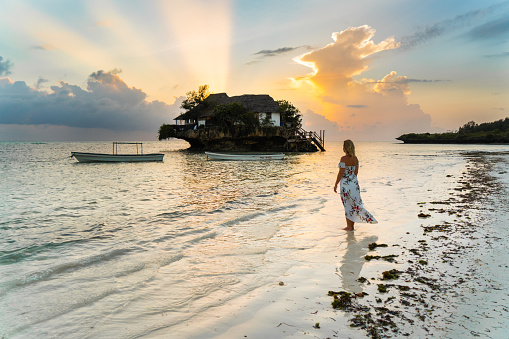 Blond female tourist in a sundress standing on a beach and looking at the view of a house on a small island under a moody sky at sunset,  Zanzibar,  Tanzania