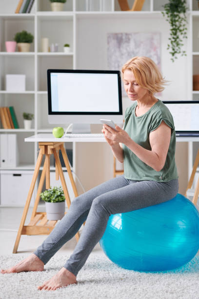 donna piuttosto matura in activewear seduta su palla fitness e usando lo smartphone - yoga business women living room foto e immagini stock