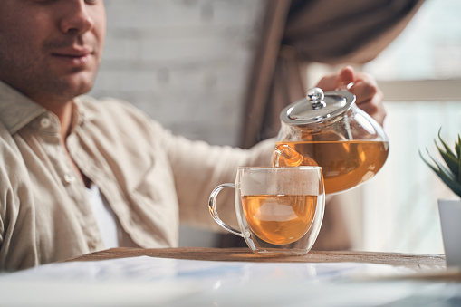 Cropped photo of a man pouring a herbal infusion into a transparent glass tea cup