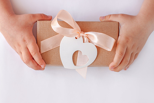 Child's hands hold gift box with ribbon bow and heart card tag. Brown kraft paper packaging flat lay. Donation concept, Giving tuesday,love holidays Valentines Day.Minimalism creative style,copy space