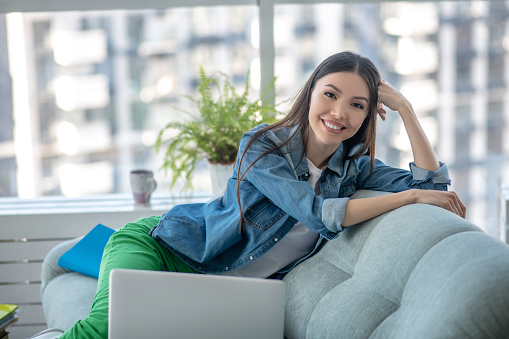 At home. Young woman in a jeans jacket sitting on the sofa and smiling nicely