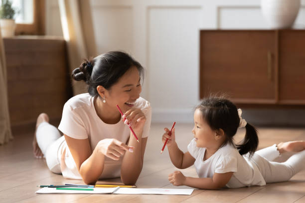 Asian mother and daughter lying on floor draw with pencils Educational pastime develop creativity skill in kid concept. Asian mother her small daughter lying on warm wooden floor in sunny cozy living room, mom teach girl paint use album and colourful pencils asian nanny stock pictures, royalty-free photos & images